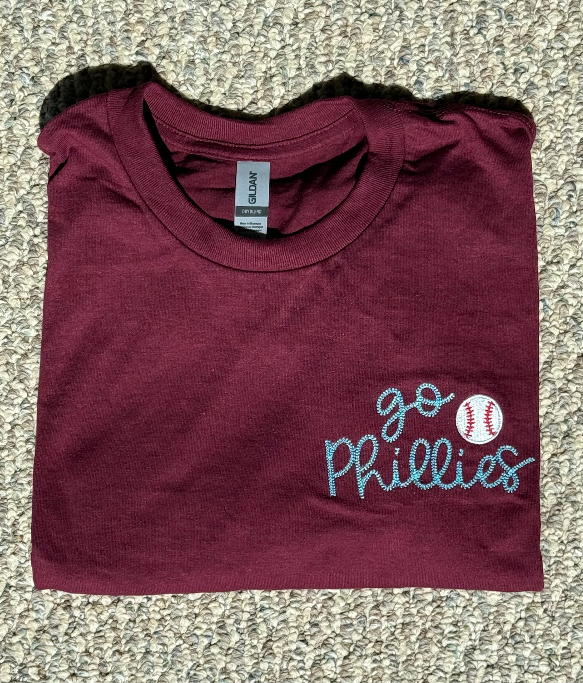 Folded maroon t-shirt with 'go Phillies' embroidered text and baseball graphic on a gray background