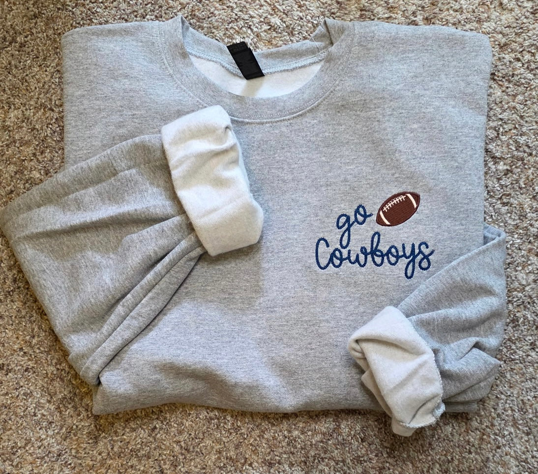 Folded gray sweatshirt with 'go Cowboys' embroidered text and football graphic on a beige background